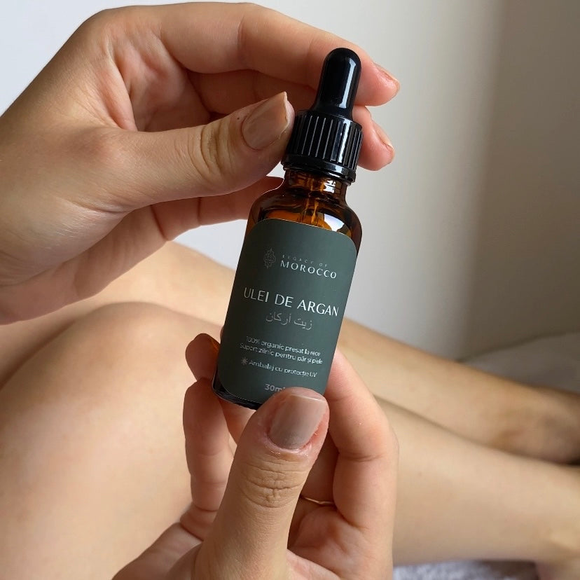 Imaginea prezintă mâinile unei persoane care ține un flacon de ulei de argan. Flaconul este din sticlă maro cu un etichetă verde pe care scrie "ULEI DE ARGAN" și alte detalii în limbi diferite, inclusiv arabă. Flaconul are o pipetă neagră ca dispozitiv de dozare. Fundalul este neutru și focalizat pe flacon și mâini.