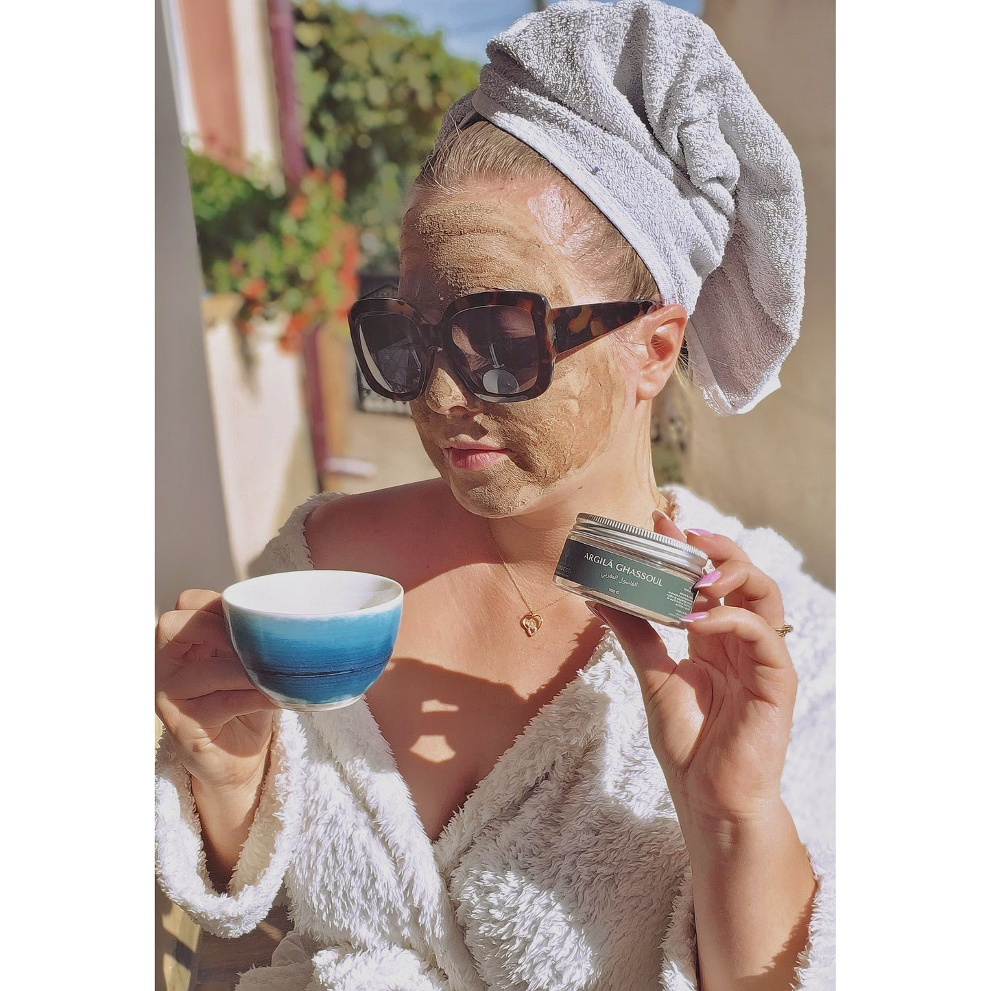 Fotografia captează o femeie în halat de baie alb, cu un prosop înfășurat pe cap, purtând ochelari de soare mari, și având aplicată o mască de argilă pe față. Ea ține o ceașcă de cafea sau ceai într-o mână și un recipient de 'ARGILĂ GHASSOUL' în cealaltă. Scena se desfășoară pe o terasă luminoasă, sub razele soarelui, sugerând o dimineață relaxantă sau un ritual de înfrumusețare în aer liber."