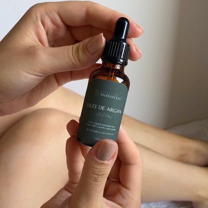 Imaginea prezintă mâinile unei persoane care ține un flacon de ulei de argan. Flaconul este din sticlă maro cu un etichetă verde pe care scrie "ULEI DE ARGAN" și alte detalii în limbi diferite, inclusiv arabă. Flaconul are o pipetă neagră ca dispozitiv de dozare. Fundalul este neutru și focalizat pe flacon și mâini.